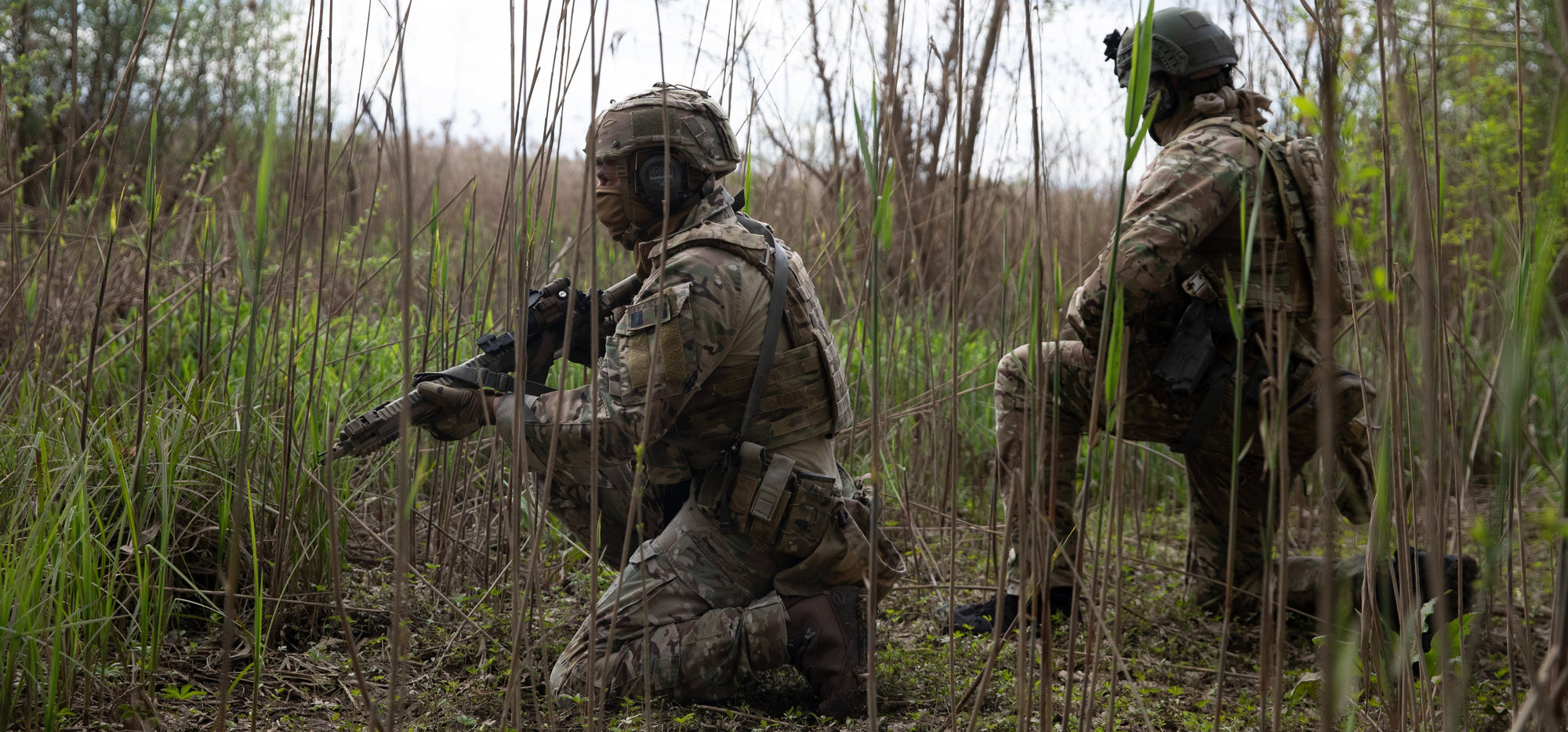 Two soldiers in camouflage gear in a grassy field wearing brown boots