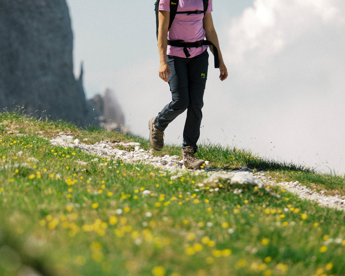 Person hiking on a grassy path with flowers and a mountain in the background